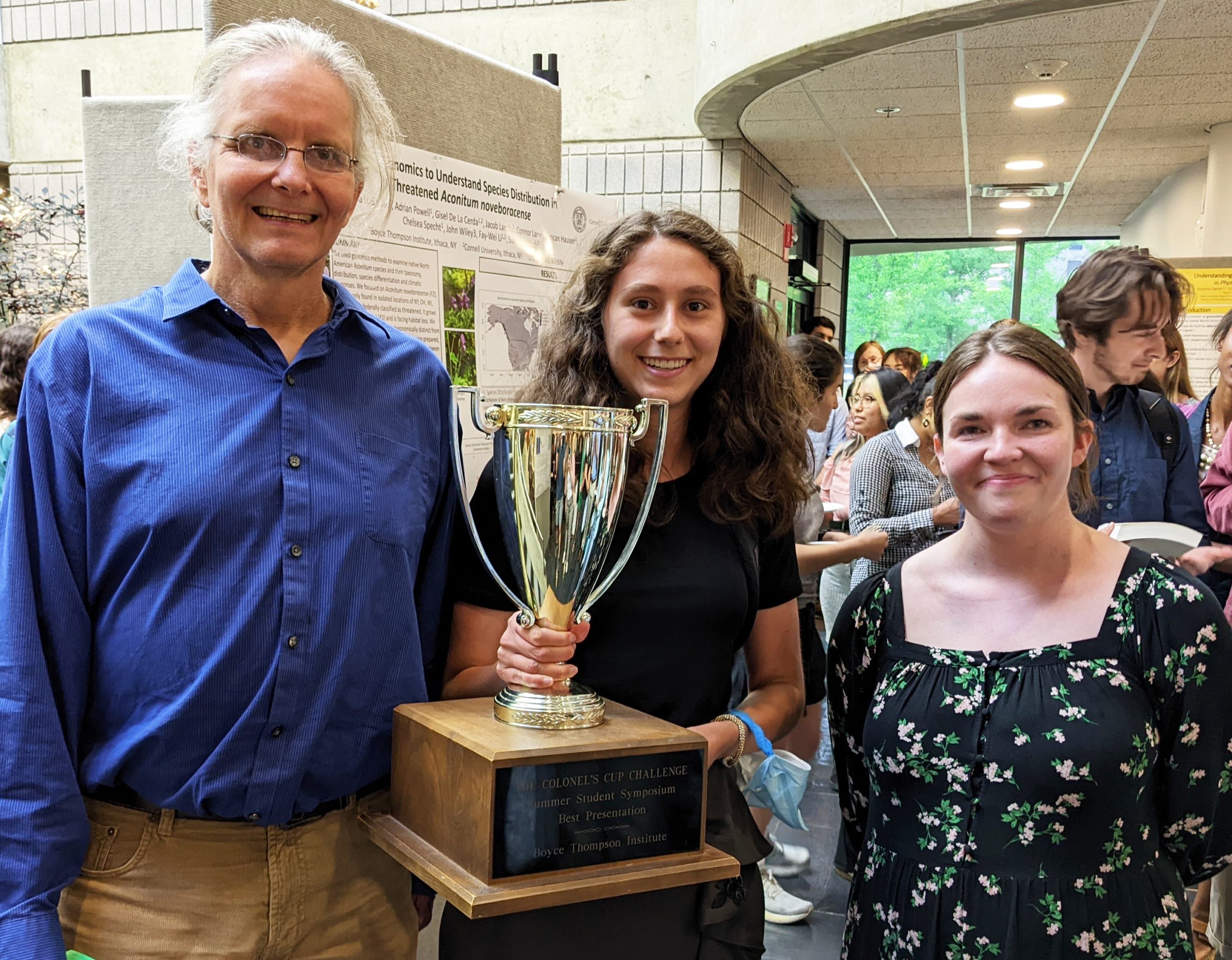 A medium shot of Georg Jander, Iselle Barrios holding a trophy, and Megan Truesdail in the BTI atrium surrounded by interns.