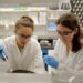 Two scientists wearing lab coats, safety glasses, and blue gloves working together in a laboratory. One is using a micropipette while the other is preparing a sample. Shelves with lab equipment and supplies are visible in the background