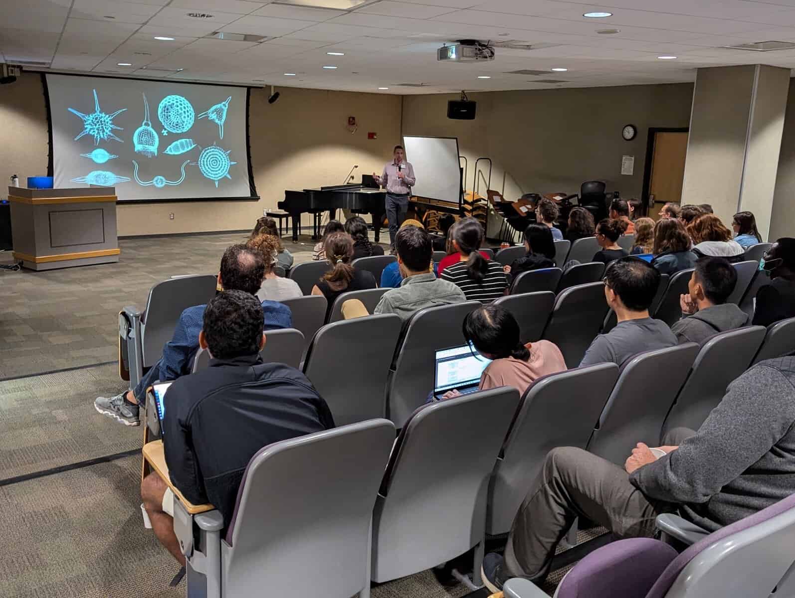 A person standing in front of a screen lecturing to a room full of people at the 2025 BTI PGS Symposium.