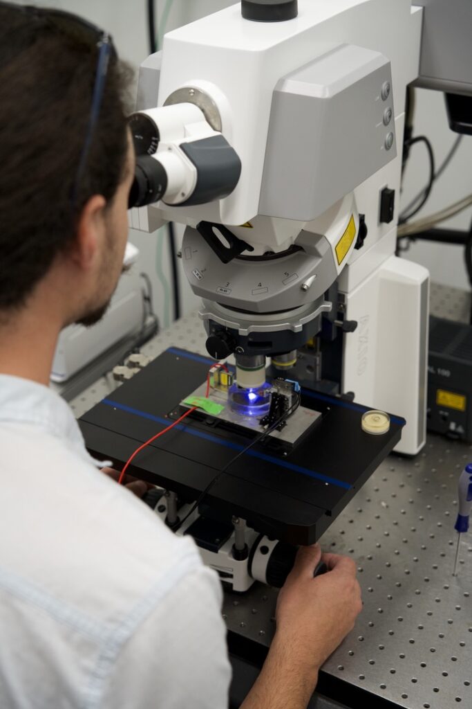 A researcher uses a high-powered microscope in a laboratory, focusing on a sample illuminated by a blue light on the stage. Wires are connected to the setup for data collection.