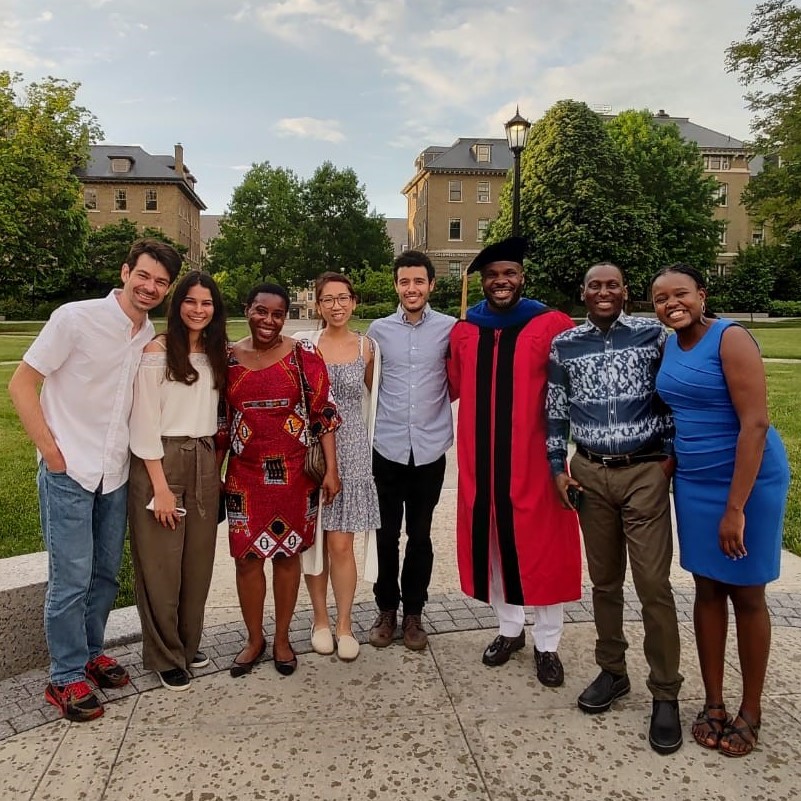 Alex Ogbonna, in his cap and gown, stands with friends and family outside on the Cornell University campus.
