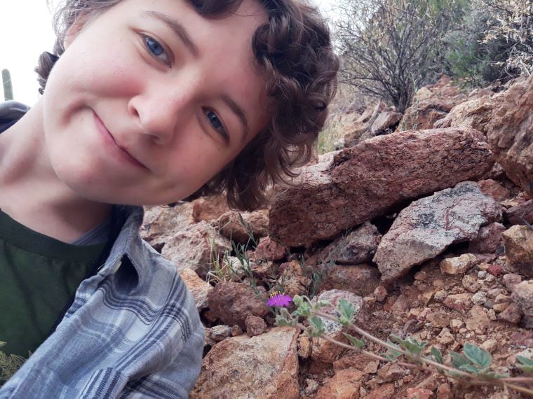 Closeup photo of Emily Humphreys with a purple flower on what appears to be a rocky hillside.