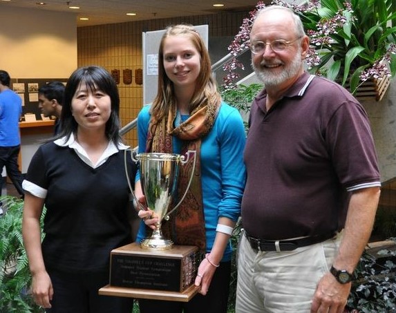 Medium shot of Dan Klessig, Miaying Tian, and Sarah Jenson smiling in the BTI atrium. Sarah is holding a large trophy.