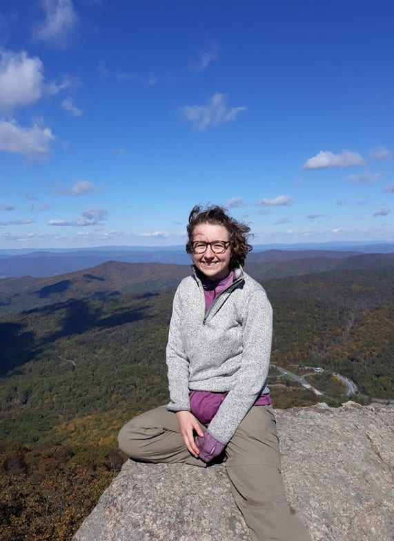 A full photo of BTI summer intern Emily Humphreys sitting on a rocky outrcop in Shenandoah National Park. The sky is blue with a few puffy clouds, and there are mountains in the distance.
