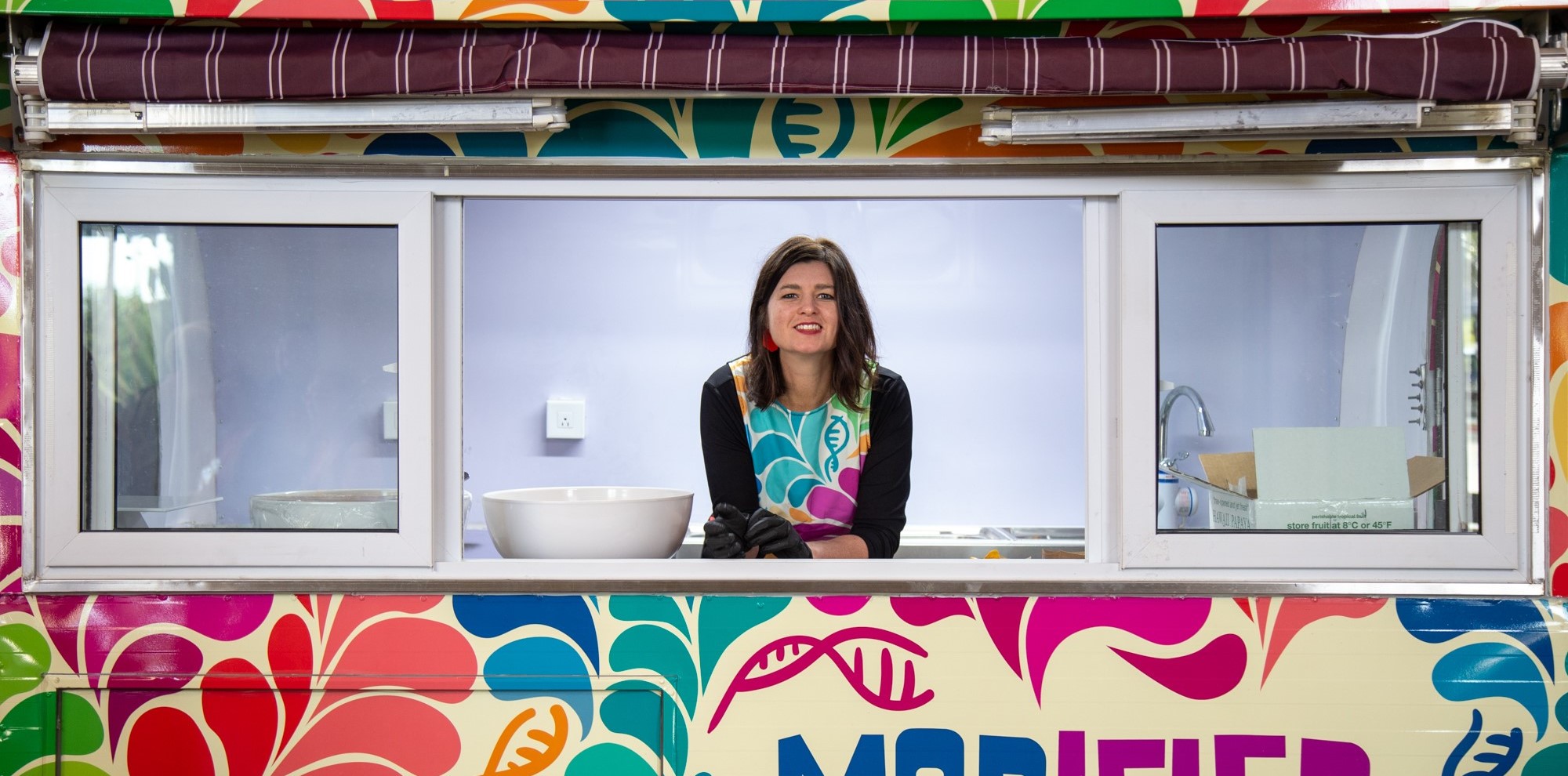Sarah Evanega smiles out of the window of a food truck. Her dress is brightly colored and matches the side of the truck, which says "Modified."