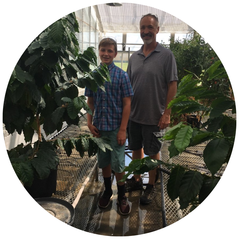Coralie Salesse-Smith and David Stern are smiling in a greenhouse full of corn plants. Salesse-Smith is reaching out and touching a plant with both hands while Stern watches.