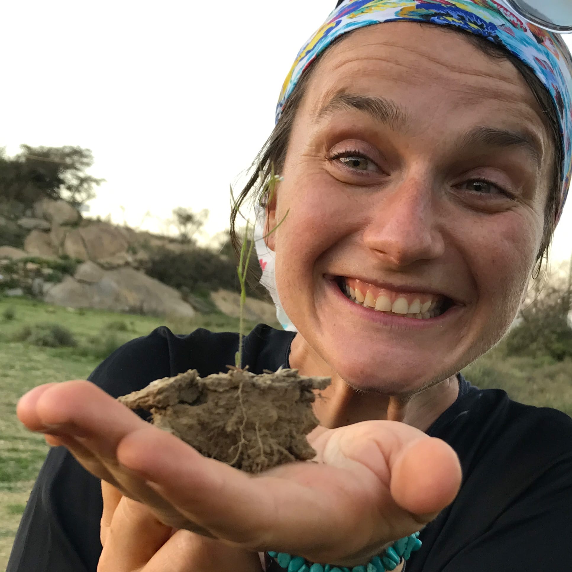 A medium closeup picture of Magda Julkowska holding a small plant in her left hand. The plant's roots are still in a clump of dirt in a way so that you can see the natural root architecture.