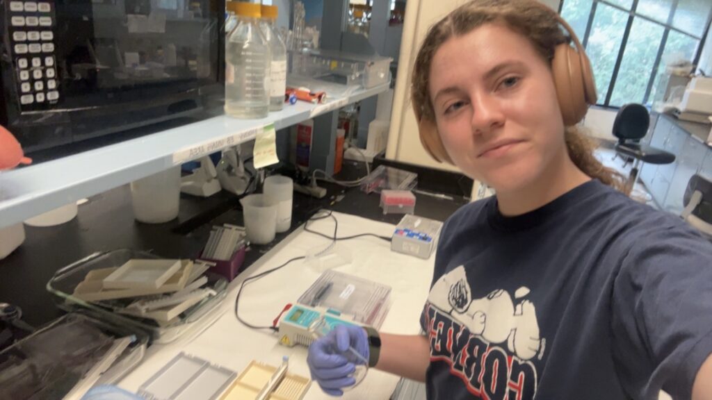 person wearing gloves and headphones taking a selfie at a lab bench with scientific equipment and supplies in the background.