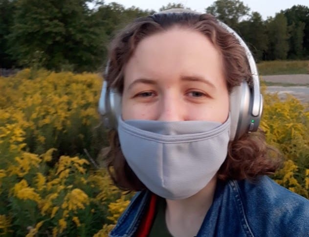 A close-up selfie of Emily Humphreys wearing a gray mask and over-the-ear headphones in a field of goldenrod.
