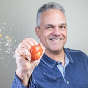 Close up of Jim Giovannoni squishing a red tomato with juice flying out towards the camera