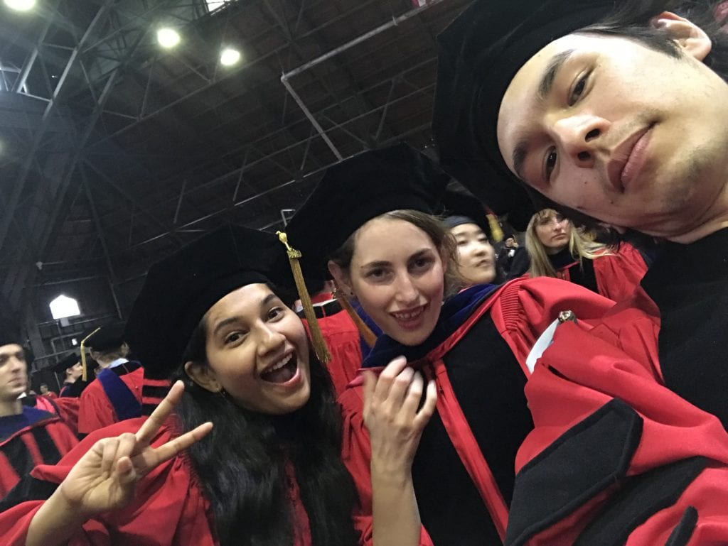 Pooja, Penelope, and Maro wearing their capes and gowns celebrating their accomplishment by taking a selfie.