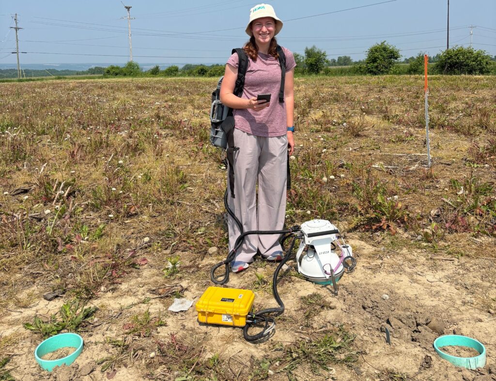 A student researcher stands in a field on a sunny day, wearing a hat, backpack, and light-colored clothing. They are smiling and holding equipment used for soil or plant measurements, including a white sensor device with cables connected to a yellow case on the ground. The field is patchy with plants, and power lines stretch across the background.