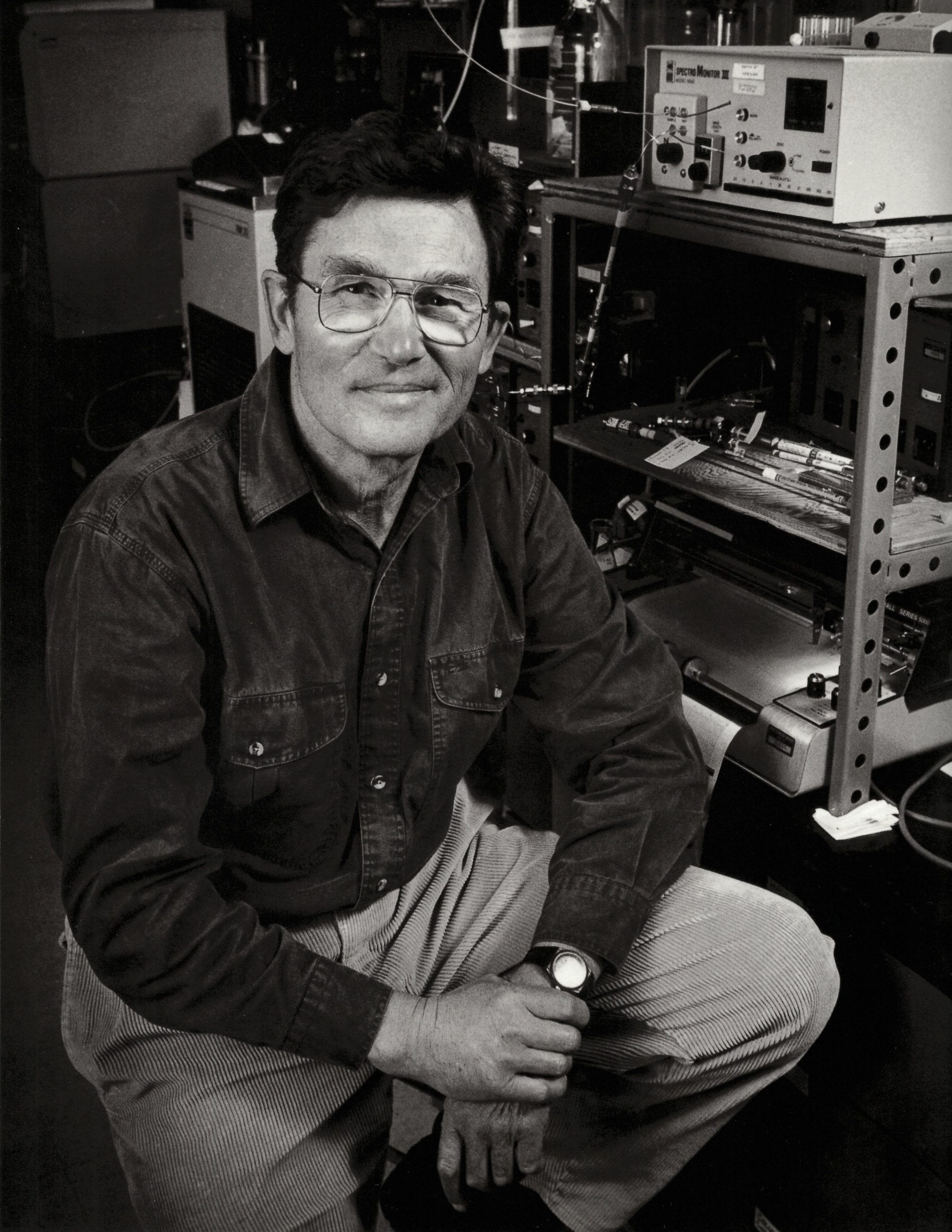 A black and white photo of Vlado Macko sitting on a stoll in front of some scientific equipment.