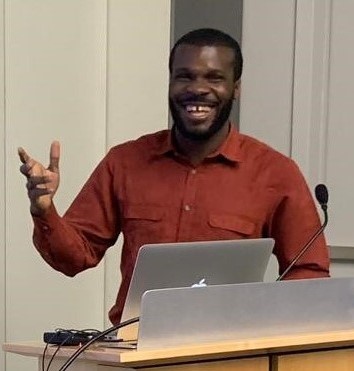 Alex Ogbonna stands behind a lecture with a big smile. He is wearing a long-sleeved, button-up, collared shirt that is reddish-orange. He is gesturing with his right hand.